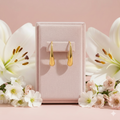 Gold earrings in a pink jewelry box with white lilies and pink flowers on a pink background