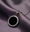 Round black pendant with silver border on a chain, placed on purple fabric.
