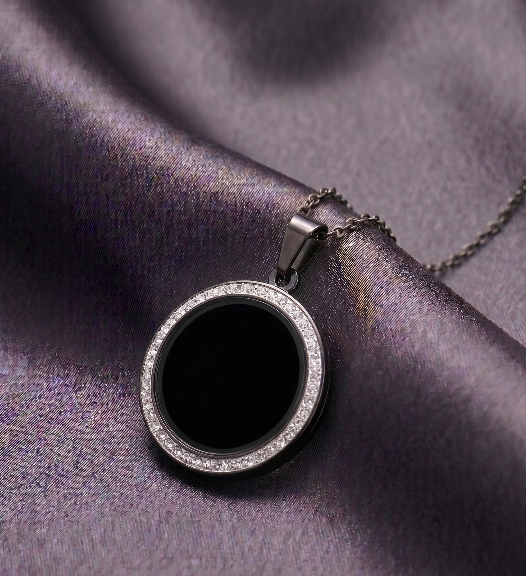 Round black pendant with silver border on a chain, placed on purple fabric.