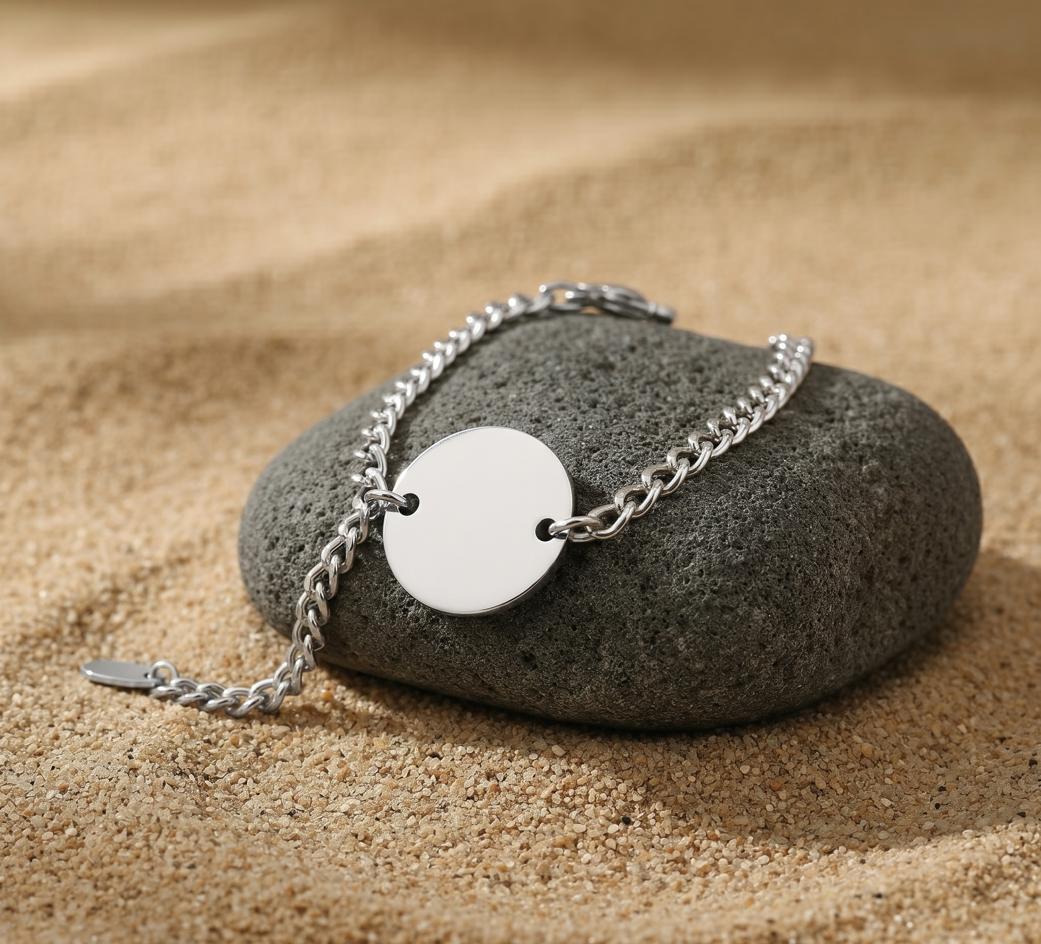Silver bracelet with a round silver charm on a dark gray stone with sand background