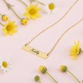 A gold bar necklace with a rectangular blank pendant on a delicate chain, displayed on a textured white surface with a curved white accent.